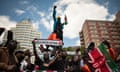 Protesters stand on a street holding a sign that says ‘Rex Masai avenue’. Some are also holding Kenyan flags and one person stands with their fist raised in the air