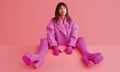 Zawe Ashton wearing a shocking pink jacket, gloves, tights and shoes, with her legs up on the table in front of her, shot against a pink backdrop