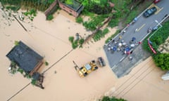 Aerial photo of people standing on the edge of a flooded street as others are carried over the water in the front bucket of a bulldozer