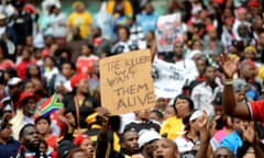 A man in a crowd at a football stadium at the funeral of Senzo Meyiwa, the national football captain, holds a sign high above his head reading "The killers, we want them alive."