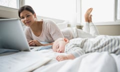 A young mother lies on her bed working on a laptop as her baby sleeps next to her