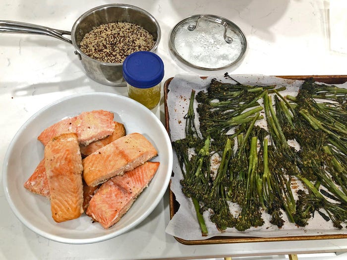 A bowl of salmon, Broccolini on a baking sheet covered in parchment paper, quinoa in a pot with the lid next to it, and a container of vinaigrette on a white countertop.