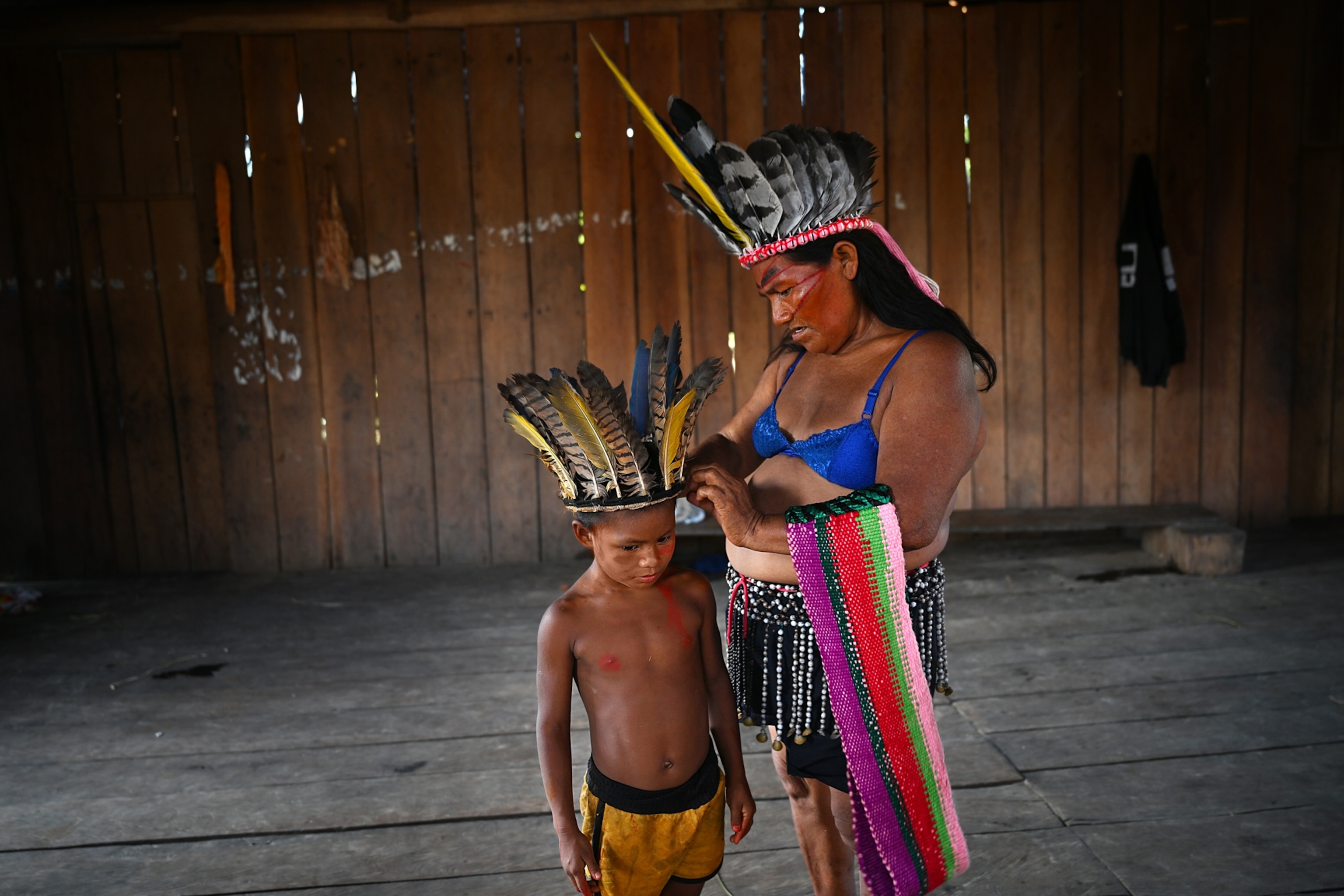 Woman in cobalt blue bra and bidded skirt with headdress of feathers affixing feather headdress on little boy.