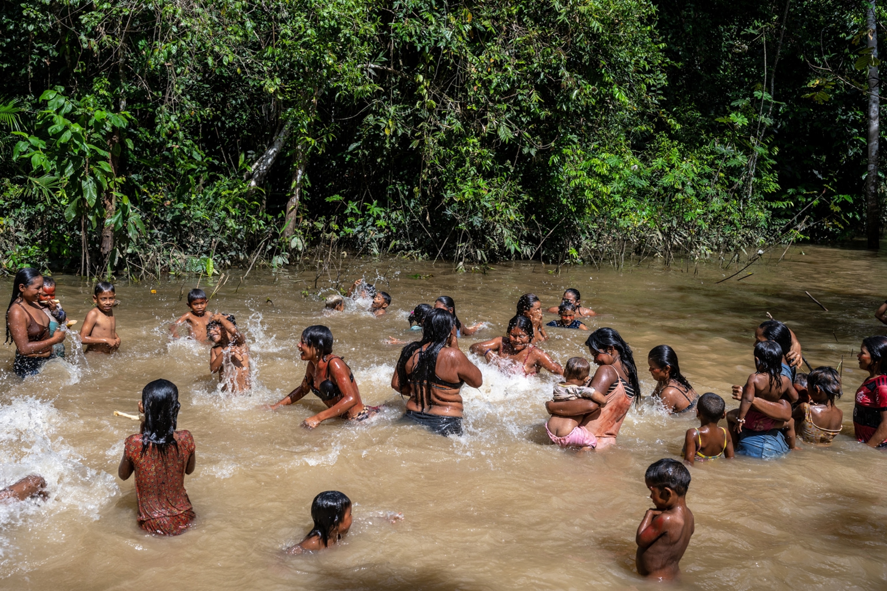 Women and children bathing in murky water by the green wall of the forest.