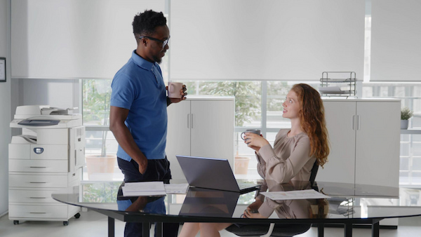 Multiethnic male and female work colleagues discussing at desk looking at laptop and drinking coffee