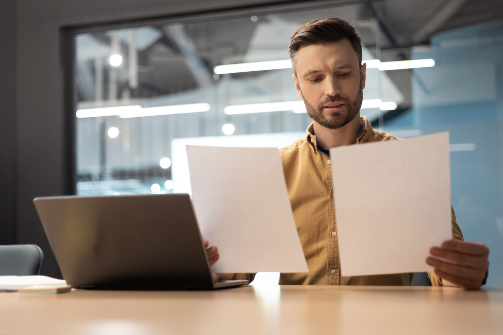 Business Man Holding Two Papers Doing Paperwork Sitting Near Computer At Workplace Indoor. Male Entrepreneur Reading Financial And Taxes Bills In Modern Office. Entrepreneurship And Business Career