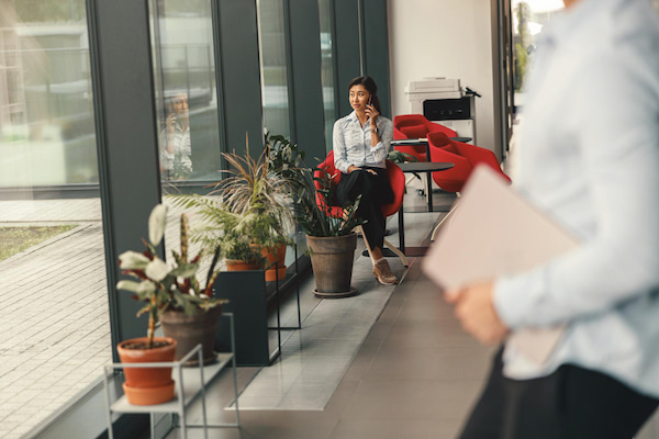 Woman office worker sitting near window and talking by phone with colleague on background