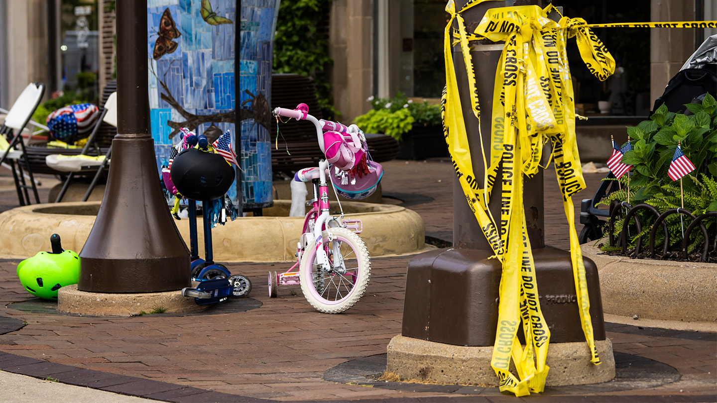 Yellow police tap wraps around a black pole. To the left are children's bikes with a green helmet abandoned on the brick sidewalk. The scene comes a day after someone used a gun to kill and injure people at a parade.