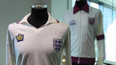 Viv Anderson's England shirt from his international debut in 1978. It is white with long sleeves and has several marks and some yellowing from age. An England crest is stitched on to the left breast with an Admiral manufacturer's logo on the opposite side. Behind is the white tracksuit top he also wore that day. It has an England badge, Admiral logo and red and blue stripes on the collar, sleeves and hem.