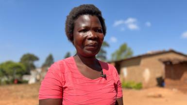 Dorothy Masasa standing outside in a pink t-shirt in the sun. There is a single storey building in the background.