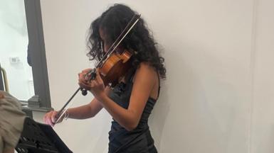 A woman plays a violin. She is wearing a dark dress and has long curly hair which is obscuring her face