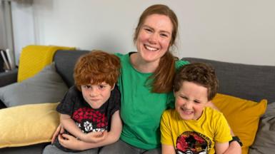 Cara Carey sits on a sofa in a green top with her two sons on either side, smiling at the camera 
