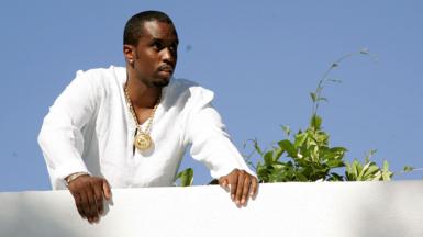 Sean "Diddy" Combs looks out over a white balcony wall while wearing a white top and gold chain, with a plant next to him and blue sky in the background, at one of his famous White Parties at his East Hapton estate in New York in 2007