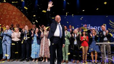 Liberal Democrat leader Sir Ed Davey waves after delivering his keynote speech at Liberal Democrat Party Conference in Brighton, with confetti and rows of MPs clapping behind him.