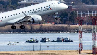 An passenger jet takes off with the wreckage of another passenger jet in the river in the background.