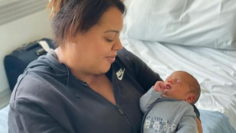 A woman with dark hair tied back in a ponytail, wearing a navy blue hoodie, looks down at her sleeping baby with a smile - they are sat on a bed, with a suitcase visible behind them 