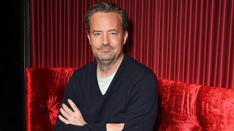 Matthew Perry looks at the camera and crosses his arms against a red background 