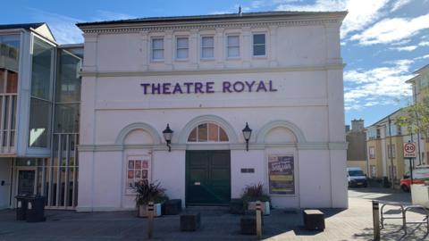 A theatre building with plants outside and an array of other street furniture