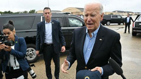 Biden gestures while speaking to reporters on the tarmac at Joint Base Andrews in Maryland on 2 October, with a security official, black SUVs and a reporter with a camera in the background.