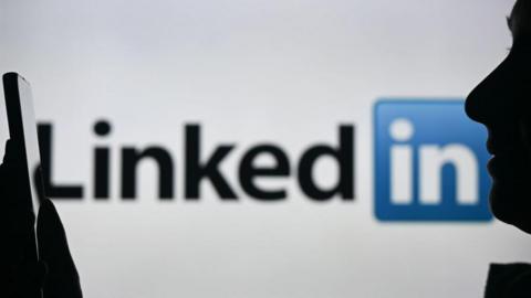 A silhouette of a woman holding a smartphone in front of a LinkedIn logo displayed on a computer screen behind her