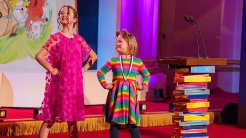 Two young girls, 500 Words participants, smiling on a stage next to a colourful backdrop and stacked books style lectern with microphones.