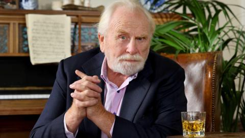 A man with a beard dressed in a suit jacket and pink shirt sits in front of a piano with a glass of whisky at his hand