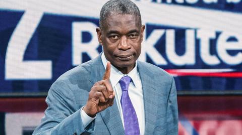 Dikembe Mutombo, wearing a light blue suit with a purple tie, raises an index finger towards the camera