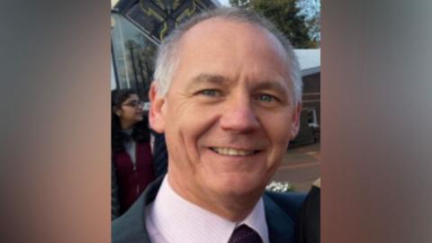 A head shot of a smiling middle-aged man wearing a shirt and tie and a jacket 