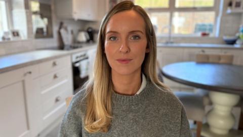 a woman with blonde hair and a grey jumper, in a kitchen