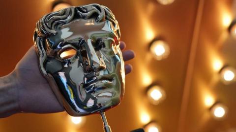 The gold mask trophy used to award BAFTA winners. It is being held up in front of a gold background with LED stage lights.