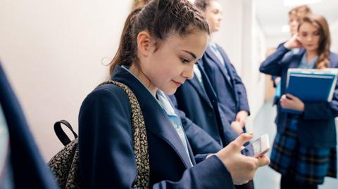 A teenage student in school uniform looks at the Bitesize app on her phone with other students in a corridor.