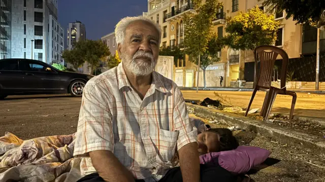 Mohamed, 55, displaced in Lebanon as a family member sleeps on the road next to him