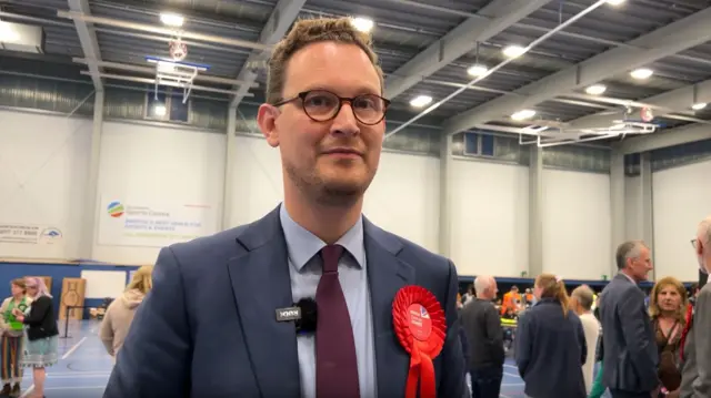 Darren Jones stood in a sports centre hall, wearing a blue suit with a red labour rosette