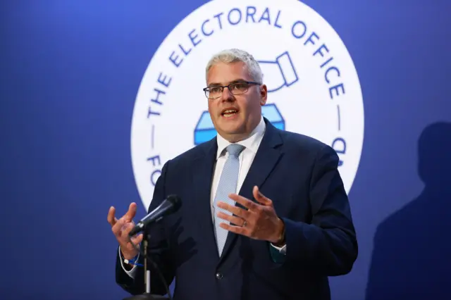 Man with white hair and glasses wearing a navy suit, white shirt and light blue tie.