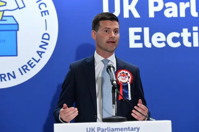 Man in blue suit with white shirt, light blue tie and red, white and blue DUP badge