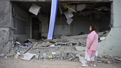 A woman wearing a pink jacket over a dress stands looking at a building that is mainly rubble