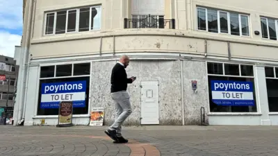 A man walks in front of an empty shop. The entrance is boarded up and  large "to let" signs, coloured blue and white, dominate two windows on the ground floor. The building is mid-20th century and has cream walls and a decorative balcony.