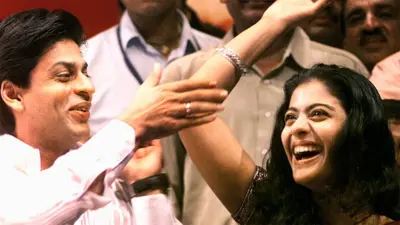 Actor Shah Rukh Khan and actress Kajol smiling and cheering, with Shah Rukh in a white shirt tapping Kajol's raised elbow