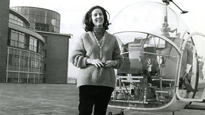 Yvonne Littlewood stands cheerfully on the roof of BBC Television Centre next to a 1960s helicopter with a round glass cockpit.