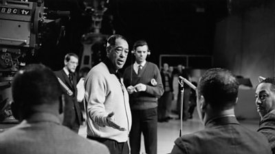 Duke Ellington gesticulates towards a seated audience as a camera looks on.