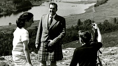 Kenneth McKellar wearing a kilt with a loch in the scene behind. Producer Yvonne Littlewood gives directions to the cameraman. 