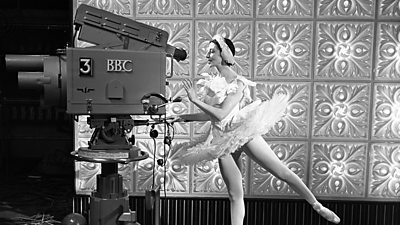 Ballerina Alicia Markova pirouetting while she poses as if operating a large BBC studio camera.