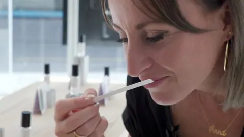 BBC Foreign correspondent Anna Holligan smells perfume. She is wearing gold earrings and a gold necklace which says Anna.