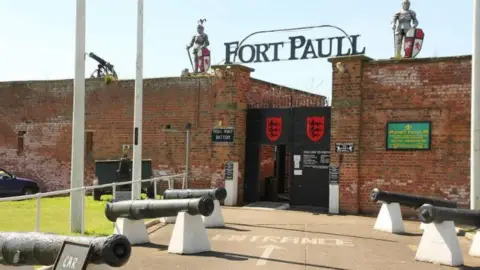 The Fort Paull entrance showing two large brick walls with a black gate in between. Two knights with shields stand next to a sign reading 'Fort Paull' on top of the wall.