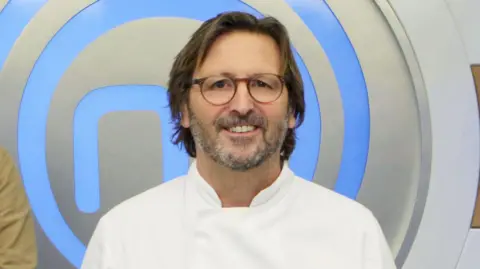 Mitch Tonks is on the set of MasterChef with a MasterChef logo behind him. He is wearing chef whites and has medium length brown hair, glasses and a grey beard.