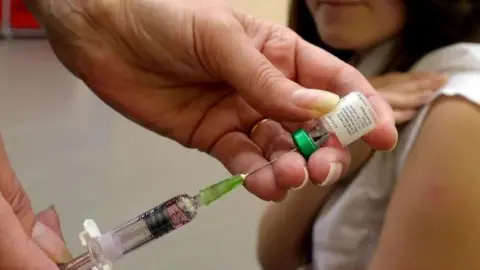 An image of two hands holding a syringe and a small glass bottle filled with liquid.  A person is in the background holding a white sleeve up at the top of their arm.