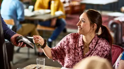 A woman sitting at a restaurant table and smiling up at a waiter as she taps her card on the contactless machine