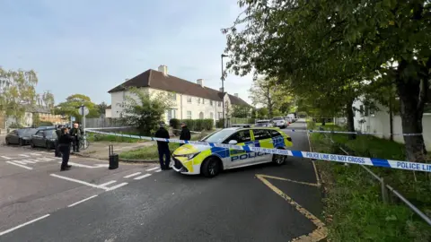 A short police cordon covering a footpath and in Waltheof Gardens housing estate. Five police officers can be seen and a police car is parked in the road as part of the cordon.