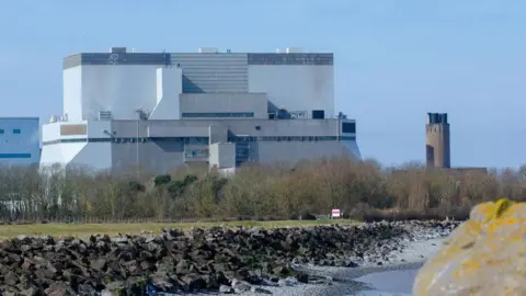 The Hinkley Point B nuclear power station, which sits near a river, in Somerset  . It also has trees surrounding it and is white building with grey trim.