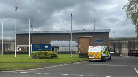 Brown, windowless building with a blue sign in front reading HM Prison Frankland and a police van parked to the right, on a grey gloomy day